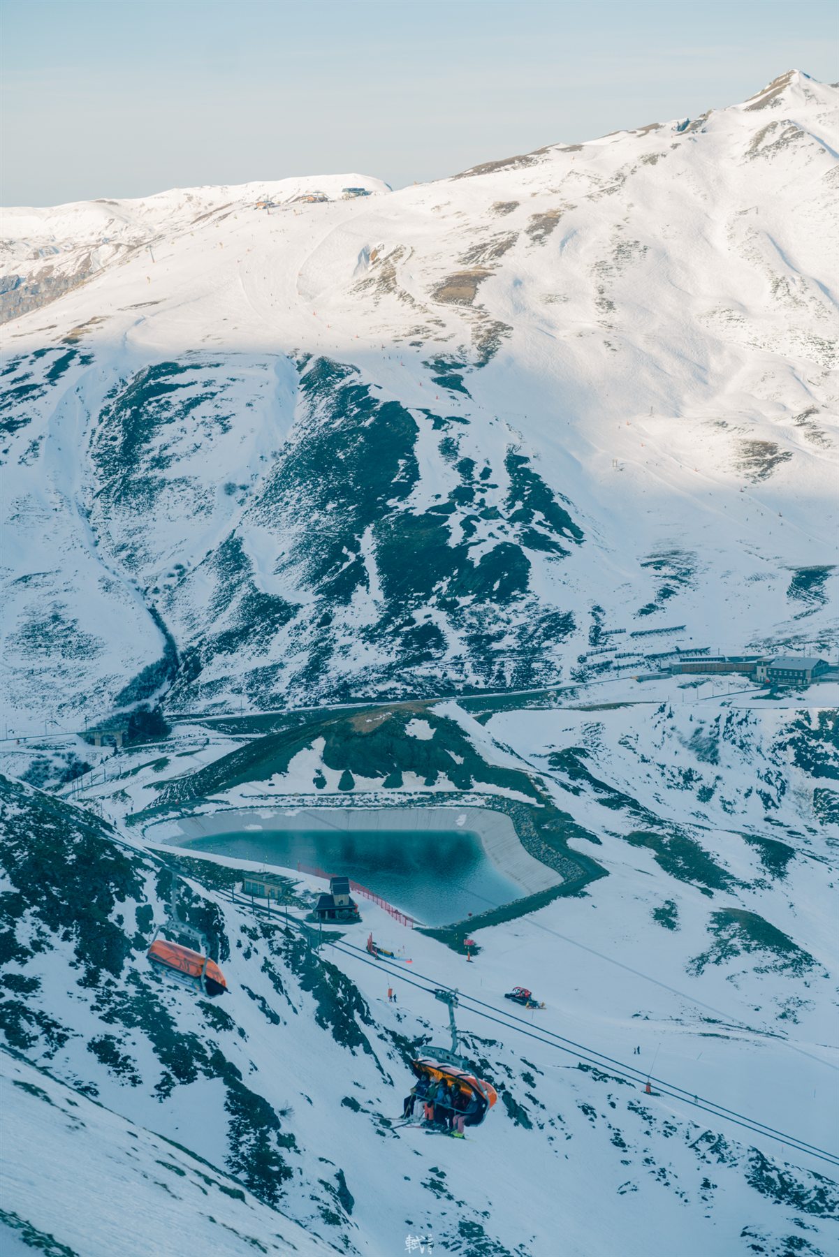 A snowy mountain slope with cable cars and a blue alpine reservoir