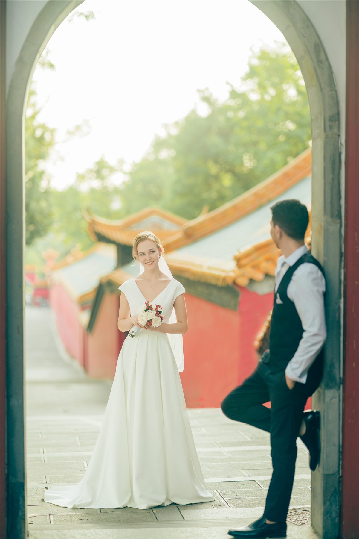 A bride and groom framed by a tall archway with red temple walls behind them